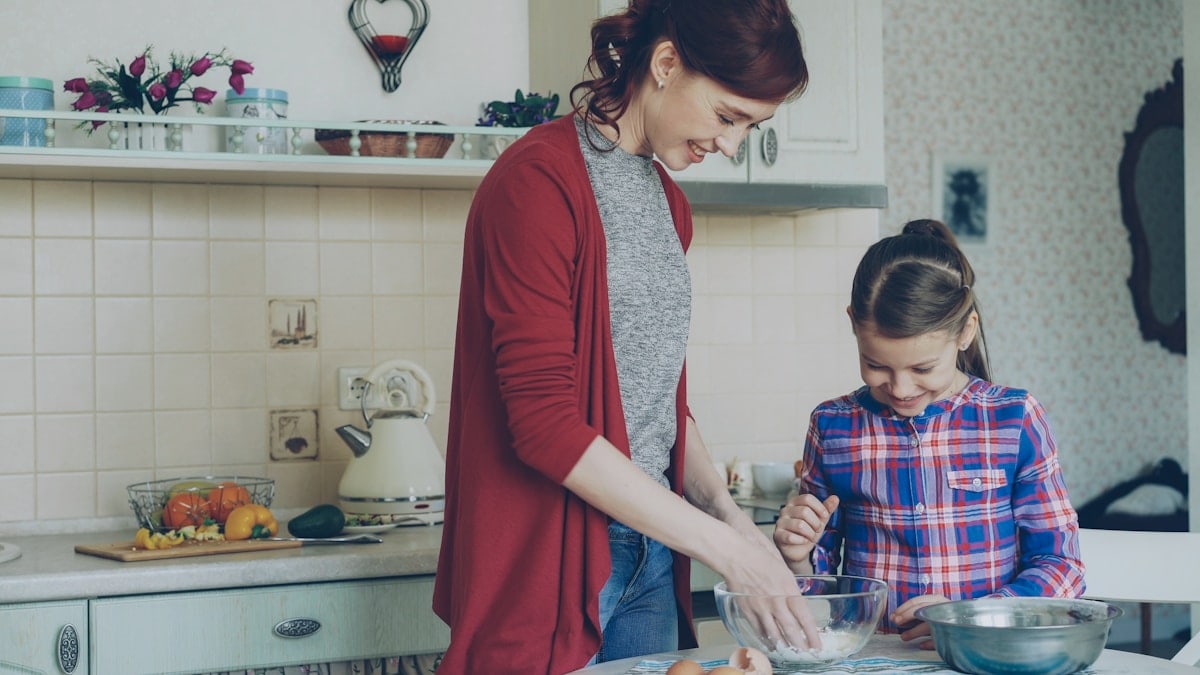 Little cheerful girl helping her mother in the kitchen mixing dough for cookies. Family, food, home and people concept